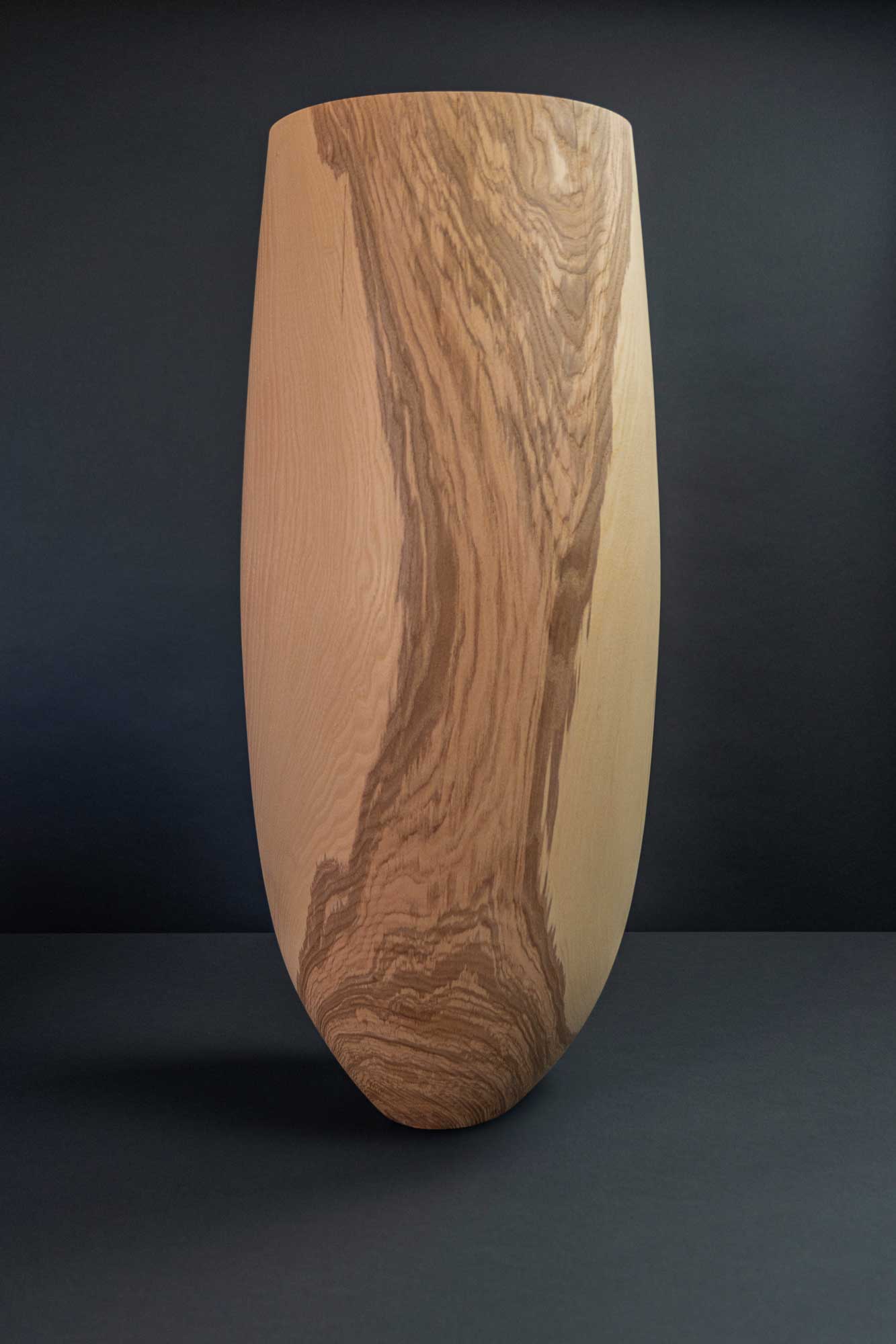 Das Bild zeigt eine große längliche Vase aus Holz vor dunklem Hintergrund.